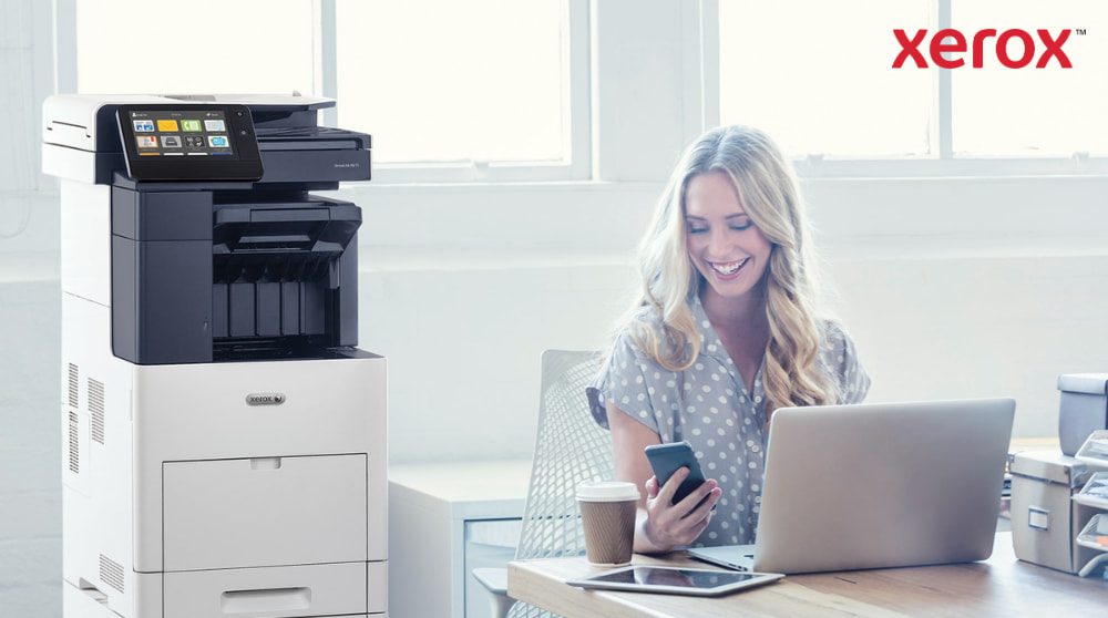 Una persona guarda felicemente il suo smartphone mentre è seduta alla scrivania con un computer portatile. Una stampante multifunzione Xerox VersaLink B605 è visibile accanto alla scrivania, in un ufficio ben illuminato.